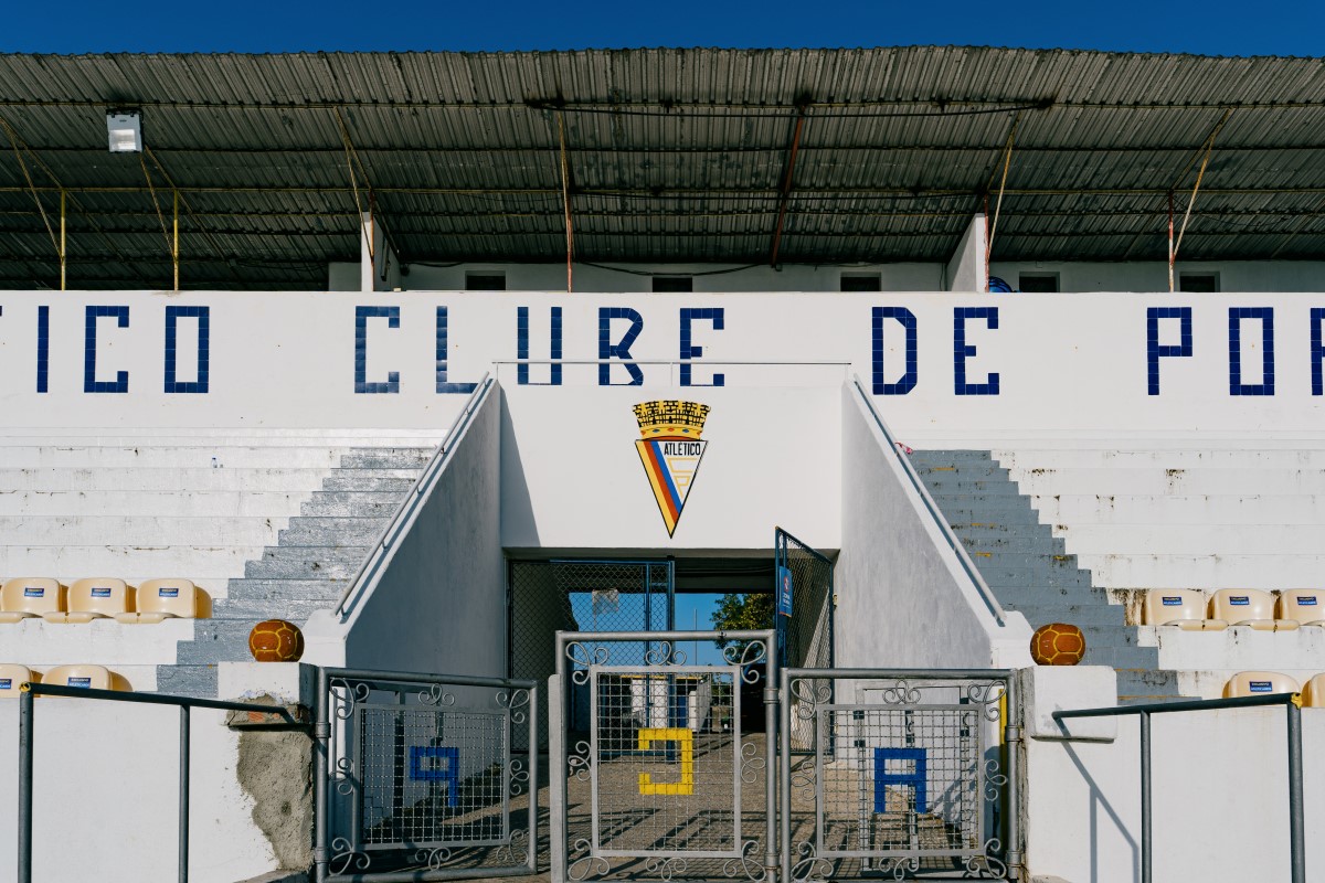 Detail of the main entrance to the stand at Tapadinha Stadium, with the emblem of Atlético Clube de Portugal at its centre. In the background, the white stands and part of the club’s name can be seen displayed in blue lettering across the façade.
