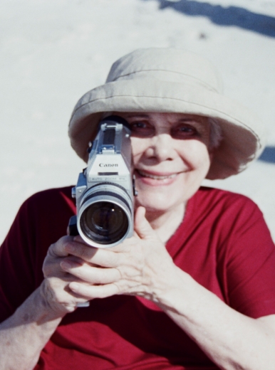 An elderly woman (Anna Maria Maiolino) in a Panama hat and red blouse aiming a Super 8 camera towards the viewer.