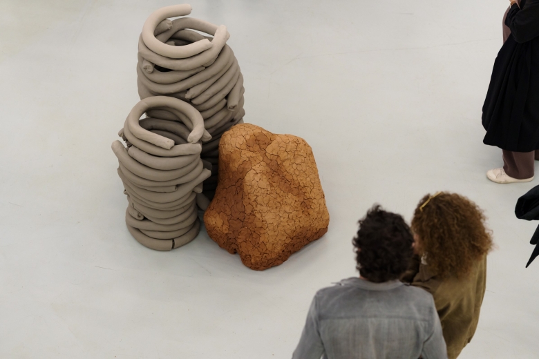 Casal contemplando uma escultura, composta por duas duas torres de tiras de argila cinzenta e uma rocha castanha