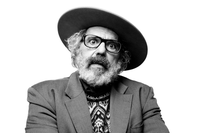 Retrato fotográfico a preto e branco de Manuel João Vieira, sentado e fotografado de um ângulo ligeiramente baixo. Usa chapéu de aba larga, óculos, casaco e camisola de malha com padrão geométrico, sobre um fundo neutro.
