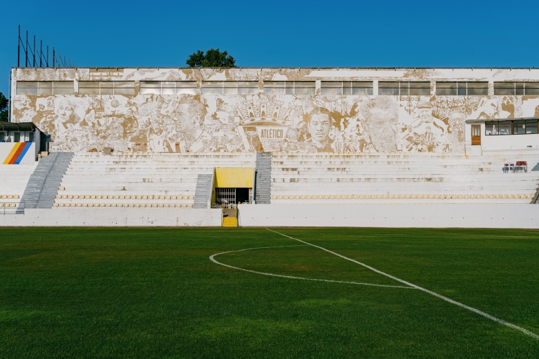 Panoramic view of Tapadinha Stadium, with Vhils’ large-scale artistic intervention Scratching the Surface Project covering the entire façade of the main stand. Viewed from the stadium pitch, the work incorporates portraits, scenes related to the history of Atlético Clube de Portugal, and the club’s emblem at its centre.