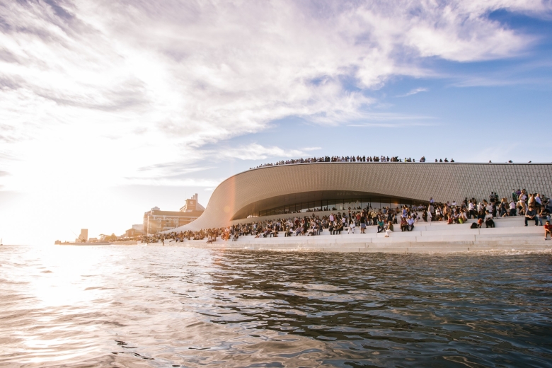 Panoramic view of MAAT in the late afternoon, with visitors spread across the undulating rooftop and riverside steps beneath a partly cloudy sky.