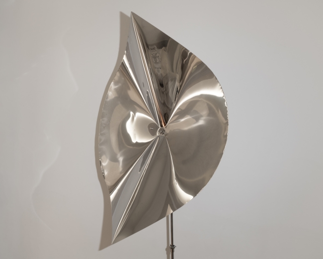 Escultura abstrata em metal polido, com formas curvas e superfície refletora, apresentada sobre um suporte diante de uma parede branca.