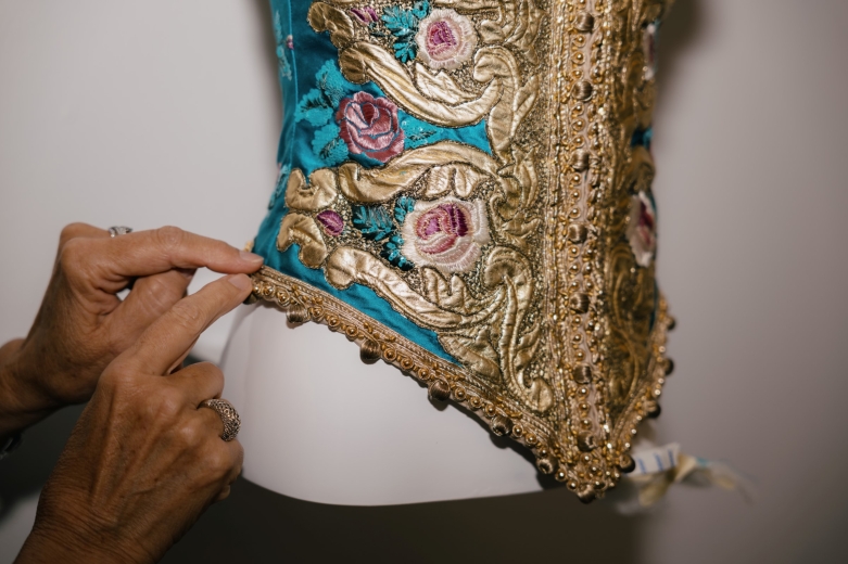 Pormenor de uma peça de vestuário em tecido azul-turquesa, ricamente decorada com bordados florais, aplicações douradas e acabamentos ornamentais. Uma pessoa segura cuidadosamente a bainha da peça para revelar os detalhes do trabalho têxtil.