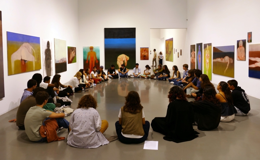A group of young people sit in a circle inside an art gallery, taking part in a guided activity surrounded by large-scale figurative paintings displayed on the walls.