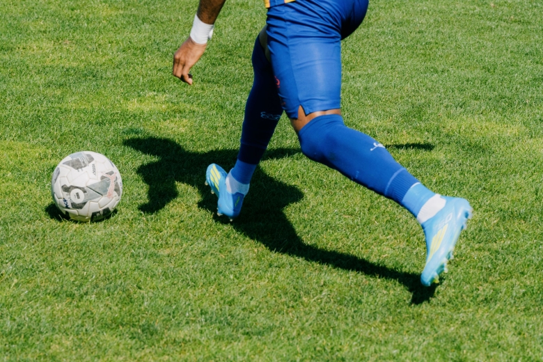 Pormenor de um jogador em movimento no relvado do Estádio da Tapadinha, aproximando-se de uma bola de futebol. A fotografia centra-se nas pernas, nas chuteiras azuis e na textura da relva.