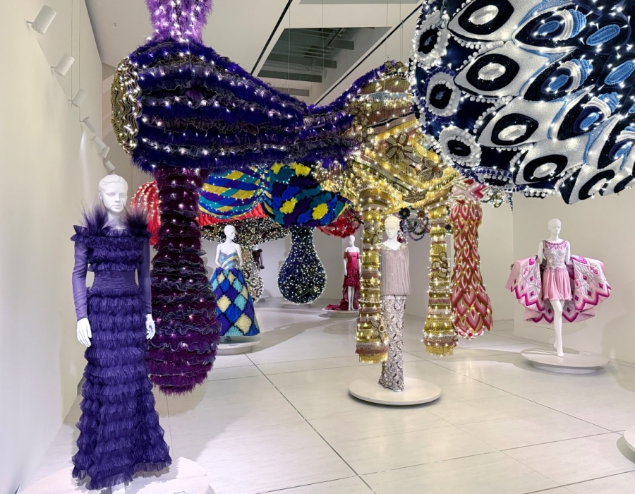 Esculturas têxteis monumentais suspensas entre manequins com vestidos de alta-costura numa galeria expositiva, criando um diálogo entre arte contemporânea e moda.