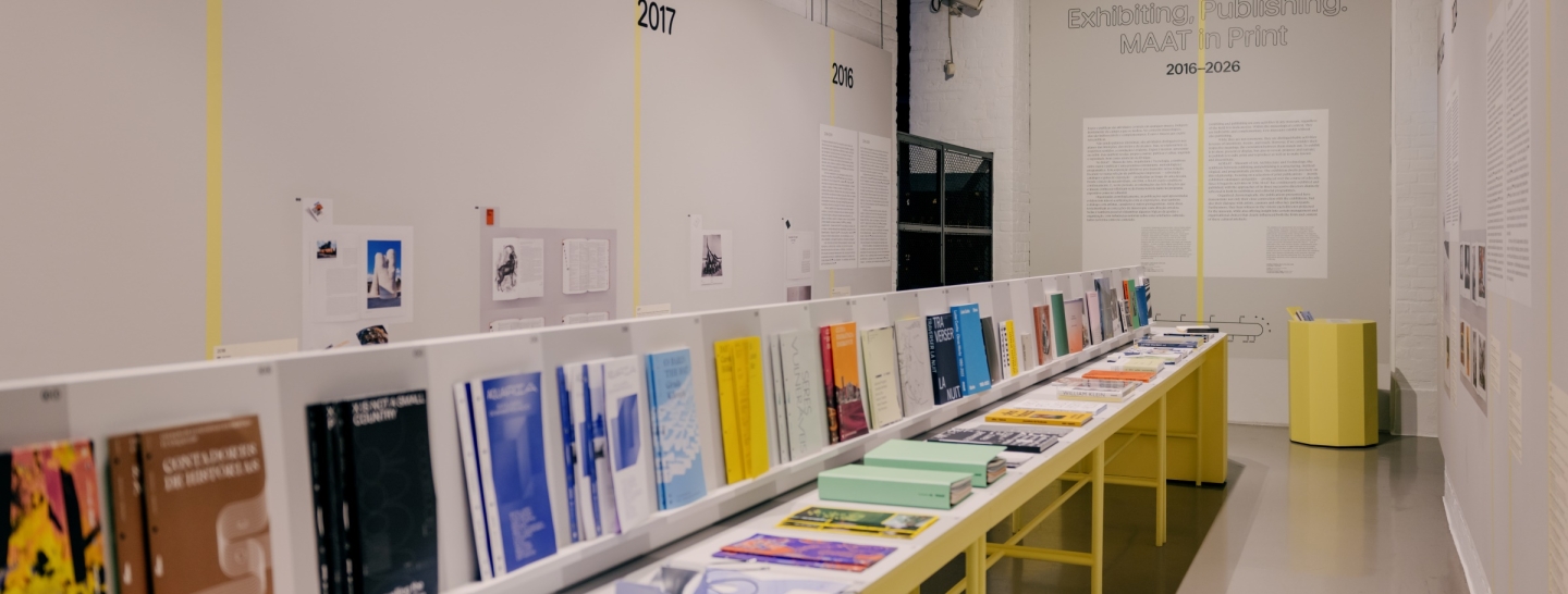 Vista de uma galeria dedicada à produção editorial do MAAT, com mesas preenchidas por catálogos, livros e publicações organizados ao longo de uma cronologia que documenta a atividade do museu entre 2016 e 2026.