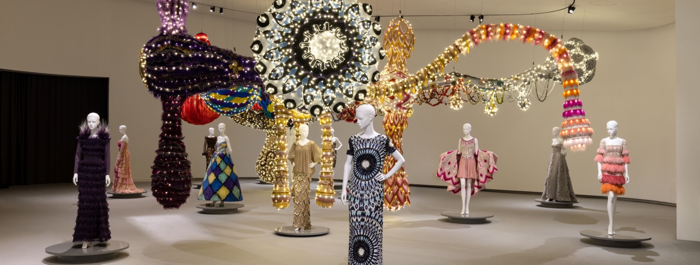 Vista geral da instalação Valquíria Vénus, de Joana Vasconcelos, suspensa sobre um conjunto de manequins vestidos com peças de alta-costura da Maison Valentino. A obra combina grandes volumes têxteis iluminados em tons de roxo, dourado, vermelho, rosa, branco e azul, ocupando o centro do espaço expositivo.